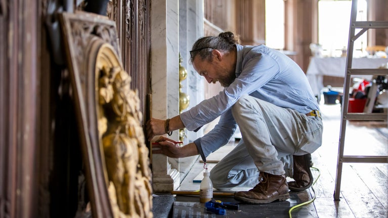 Male conservator in pale blue shirt and cream trousers kneels down to repair The Vyne's Oak Gallery paneling, with ladder to right and gold roundel on floor in foreground